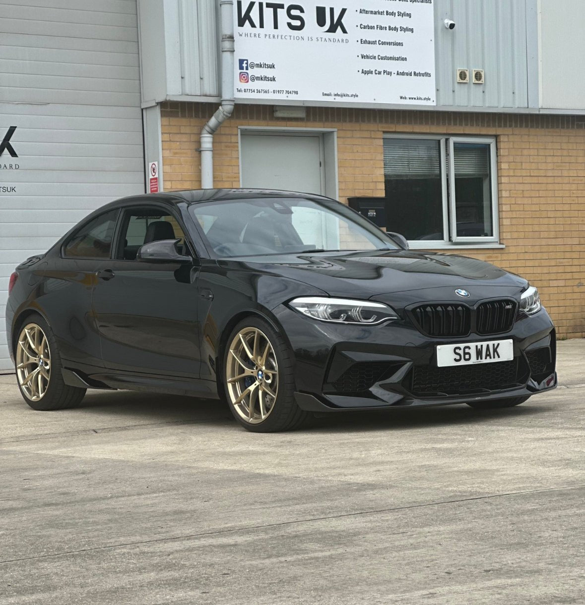 BMW M2 F87 Body Kit showcased on a black BMW M2 with gold wheels outside a garage.