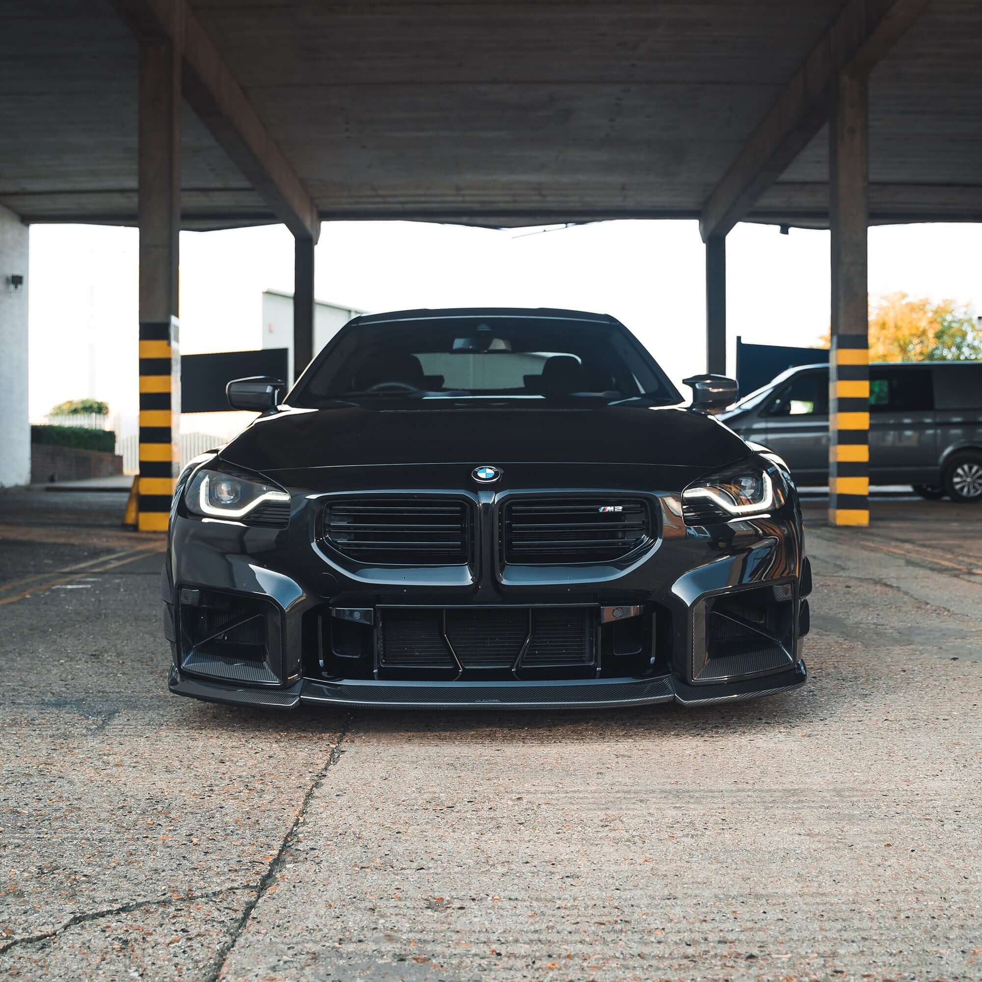 BMW G87 M2 Full Carbon Fibre Body Kit showcased on a black BMW M2 parked under a structure.