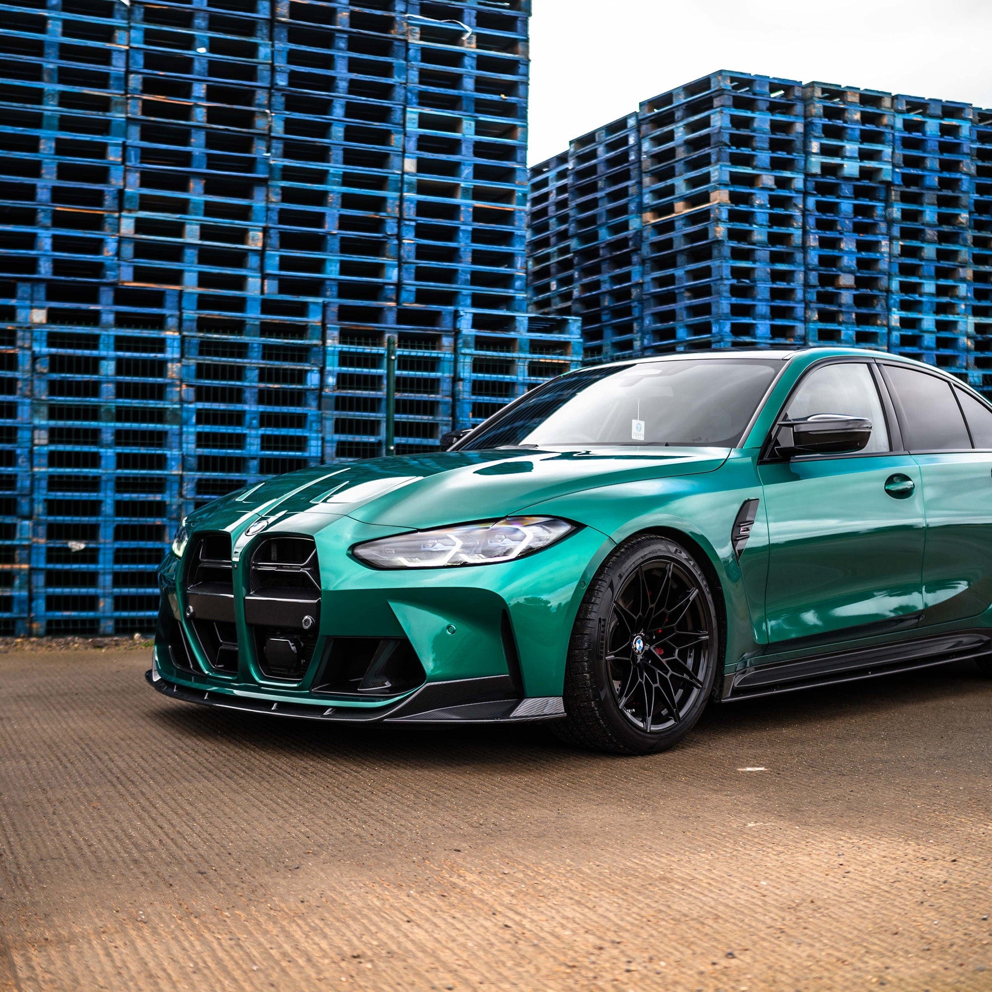 BMW G80 M3 carbon fibre splitter - V1 on a green car parked near blue pallets.