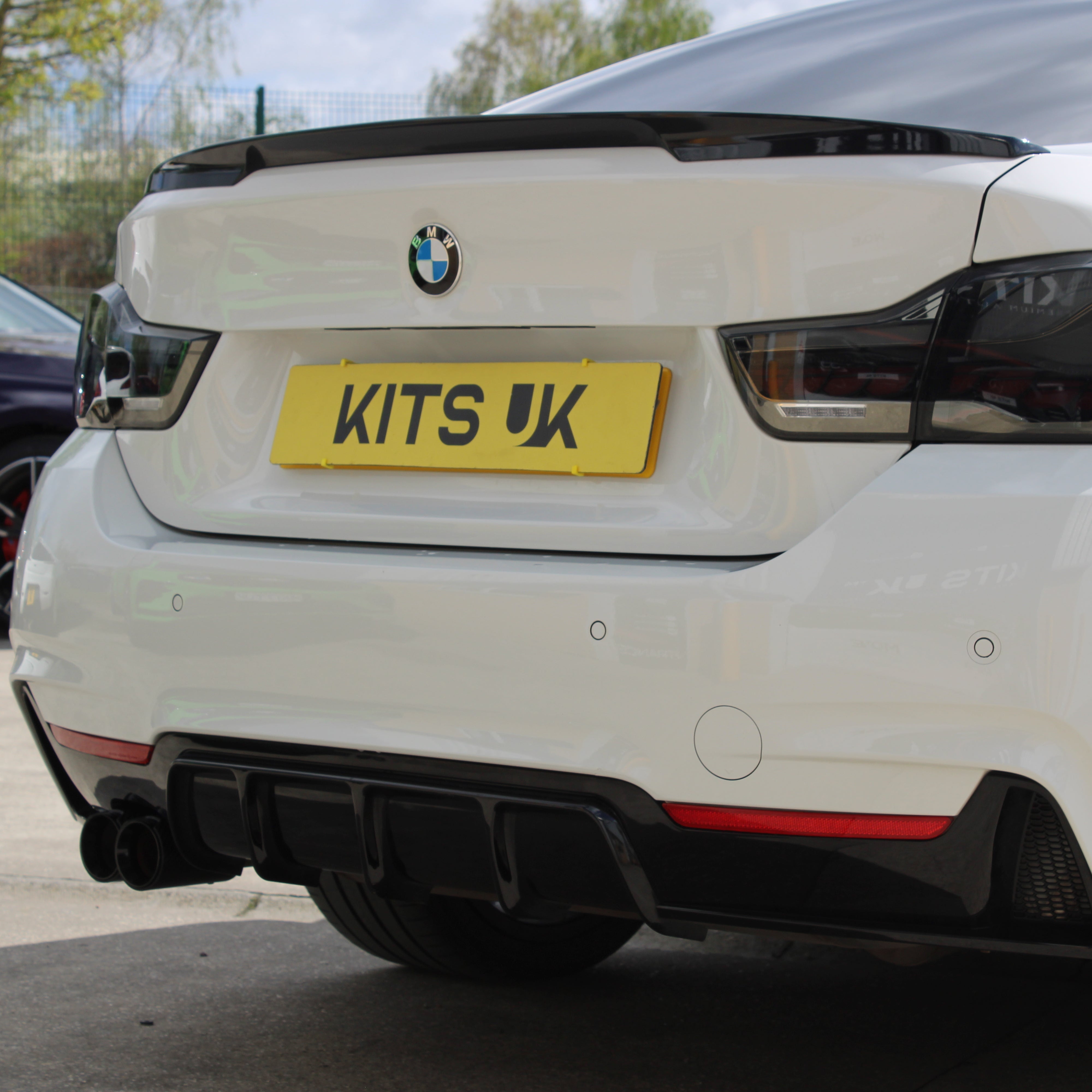 White car with a black spoiler and 'KITS UK' license plate in an outdoor setting.