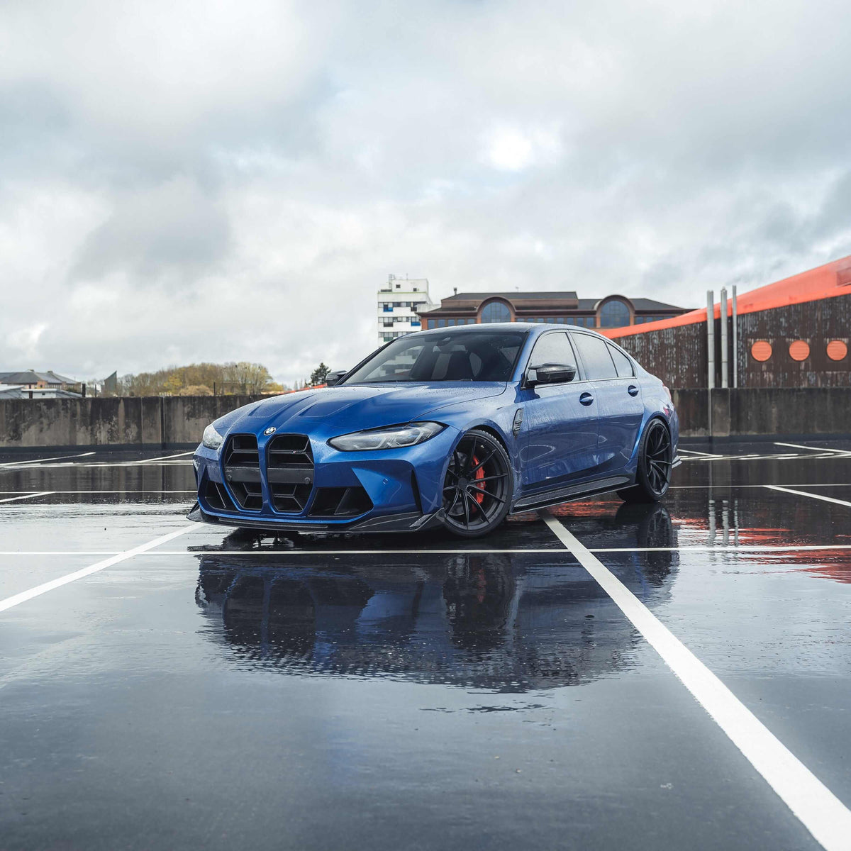 BMW M3 M4 carbon fibre splitter V2 enhancing the aggressive look of a blue BMW M3 parked on a wet surface.